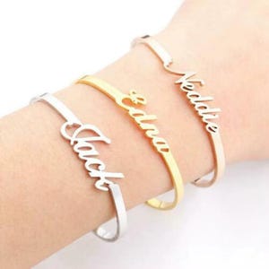 May include: Three open cuff bracelets in silver, gold, and rose gold. Each bracelet features a name in a cursive font: "Jack", "Edna", and "Veddle". The bracelets are worn on a wrist.