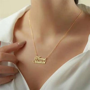 Puede incluir: Un collar dorado con los nombres "Hanna" y "Sebastian" en fuente cursiva, conectados por un diseño en forma de corazón. El collar se lleva en el cuello, contra una camisa blanca.
