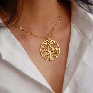Puede incluir: Collar con colgante dorado con diseño de árbol y nombres grabados en las ramas. Los nombres Lucy, Warren, Stella, Emma, Larissa y Ashley son visibles. El collar lo lleva una persona con una camisa blanca.