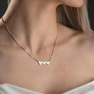Può includere: Una collana d'argento con tre ciondoli a forma di cuore. Ogni cuore è inciso con un nome: Cyrus, Dean ed Evan. La collana è indossata, con la catena e i ciondoli visibili.