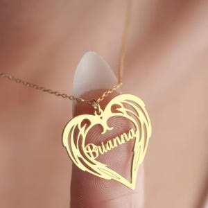 Puede incluir: Un collar dorado con un colgante en forma de coraz&oacute;n con el nombre "Brianna" inscrito. El colgante tiene un dise&ntilde;o de alas alrededor del coraz&oacute;n. El collar est&aacute; en una cadena delicada.