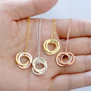 Op de afbeelding: Vier gepersonaliseerde kettingen met in elkaar grijpende ringen in goud, zilver en roségoud. Elke hanger heeft gegraveerde namen. De kettingen worden op een hand getoond, waarbij de verschillende metaalafwerkingen en ontwerpen worden getoond.
