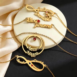 Può includere: Collane personalizzate color oro con nomi. Le collane presentano vari design, tra cui simboli dell'infinito e ciondoli circolari, ciascuno ornato di pietre preziose. I nomi sono elegantemente scritti e le collane sono esposte su una superficie bianca.