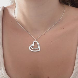 Puede incluir: Un collar plateado con un colgante de doble corazón. El colgante tiene grabados los nombres "LUKE 7.7.18" y "EMILY 6.4.20". El collar se lleva alrededor del cuello.