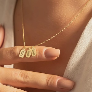 May include: A gold necklace with three rectangular name pendants. The pendants are engraved with the names "Kate", "Tom", and "Chase". The necklace is shown against a soft background, emphasizing the personalized nature of the jewellery.