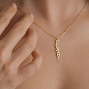 May include: A gold necklace featuring the word "PEARL" in capital letters, arranged vertically. The necklace hangs from a fine chain. The image shows the necklace worn around a person's neck.