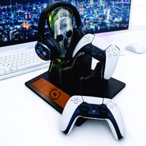 May include: A black gaming headset with red, white, and blue star accents sits on a skull-shaped stand. Two white and black game controllers are displayed with a brown leather accent piece with a "W" logo.