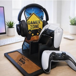 Peut inclure: Casque de jeu noir et deux manettes de jeu blanches et noires sur un support en bois noir. Le support porte un panneau Gamer Zone jaune et bleu et le texte "YOU'RE THE BEST DAD EVER".