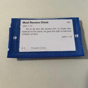 May include: A blue fabric item with white polka dots and a white rectangular label. The label contains the text "Must Receive Christ" and a Bible verse from John 1:12. The text "Proclaim Christ" is also visible.