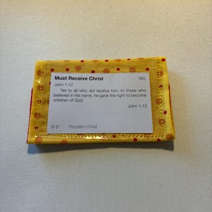 May include: A small, rectangular yellow fabric item with red polka dots and a red border. The item features text that reads "Must Receive Christ" and a Bible verse from John 1:12. The text is printed on a white rectangular label.