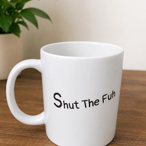 Könnte beinhalten: Ein weißer Keramikbecher mit einem abgerundeten Henkel und einer glatten Oberfläche. Auf der Außenseite befindet sich der schwarze Aufdruck "Shut The Fuh" in einer einfachen serifenlosen Schrift. Der Becher hat einen zylindrischen Körper.