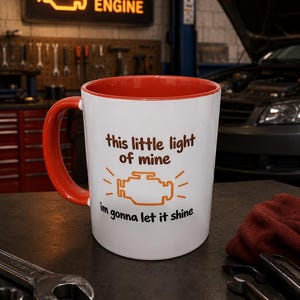 Puede incluir: Una taza de cerámica blanca con interior y asa de color naranja brillante. El frente presenta un gráfico naranja del símbolo de luz de verificación del motor con el texto "this little light of mine" arriba y "im gonna let it shine" abajo.