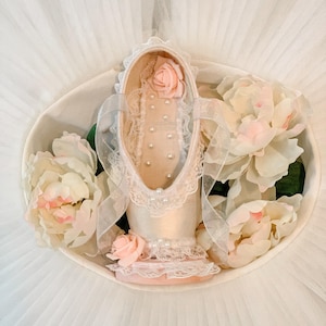 Puede incluir: Una zapatilla de ballet en un cuenco blanco, adornada con rosas rosas y perlas. El zapato es de un rosa suave con delicados adornos de encaje y cintas. El arreglo se encuentra sobre un fondo de tul blanco.