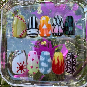 Puede incluir: Una caja de plástico transparente muestra diez uñas artificiales con varios diseños. Las uñas presentan patrones como rayas, triángulos, corazones, nubes y llamas, en colores como amarillo, naranja, rosa y verde. El fondo incluye vibrantes flores moradas.