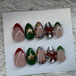 May include: A set of press-on nails with a festive Christmas theme. The nails feature designs including a red French tip, a green nail with a gold star, a nude nail with colorful lights, a black and white plaid nail with a red bow, and a green tip.