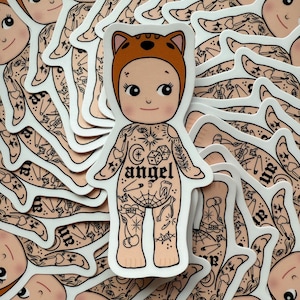 May include: A sticker featuring a cartoon baby with a cat-eared hood and tattoos. The baby has a smiling face and is covered in various tattoos, including the word "angel" and dice. The sticker is surrounded by other similar stickers.