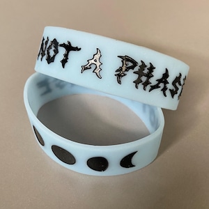 May include: Two light blue silicone wristbands. The top band has the phrase "NOT A PHASE" in black gothic lettering. The bottom band features black moon phase designs. These accessories are suitable for everyday wear.