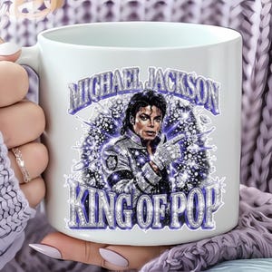 Op de afbeelding: Witte keramische mok met een afbeelding van Michael Jackson, de tekst "MICHAEL JACKSON" en "KING OF POP" in zilver en paars. De mok wordt door een persoon vastgehouden.