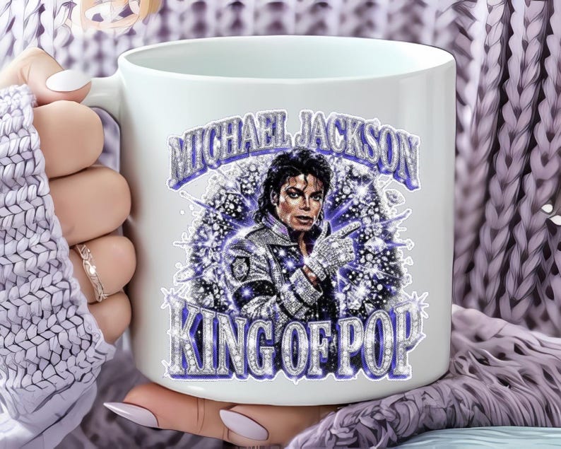 Op de afbeelding: Witte keramische mok met een afbeelding van Michael Jackson, de tekst "MICHAEL JACKSON" en "KING OF POP" in zilver en paars. De mok wordt door een persoon vastgehouden.