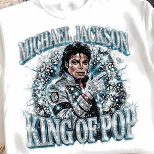 Op de afbeelding: Wit T-shirt met een afbeelding van Michael Jackson, de tekst "MICHAEL JACKSON" erboven en "KING OF POP" eronder. Het ontwerp heeft een sprankelend, juweelachtig effect met blauwe en zilveren tinten.