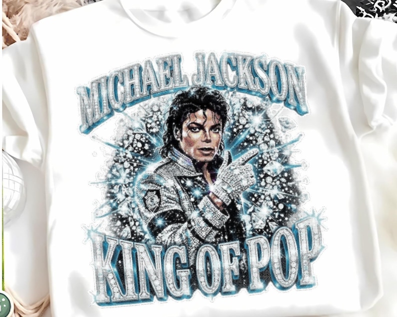 Op de afbeelding: Wit T-shirt met een afbeelding van Michael Jackson, de tekst "MICHAEL JACKSON" erboven en "KING OF POP" eronder. Het ontwerp heeft een sprankelend, juweelachtig effect met blauwe en zilveren tinten.