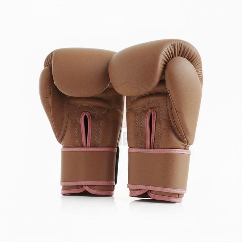 May include: A pair of brown boxing gloves with pink detailing. The gloves are made of leather and have a padded design. The wrist straps are brown with pink trim. These gloves are designed for boxing and other combat sports.