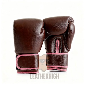 May include: A pair of brown leather boxing gloves with pink trim. The gloves feature a classic design with padded areas for protection and a secure wrist strap. The brand name "LEATHERHIGH" is printed at the bottom.