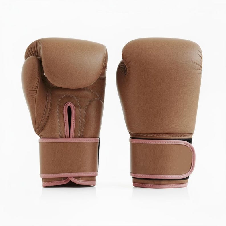 May include: A pair of brown boxing gloves with pink detailing. The gloves are made from a smooth material and have a wrist strap with a hook-and-loop fastening. These gloves are designed for boxing and other combat sports, offering protection and performance.