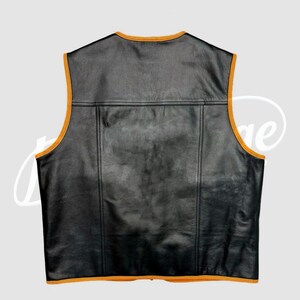 May include: Black leather vest with orange trim. The vest has a classic design with a V-neck and a rectangular panel across the upper back. The edges are trimmed with a contrasting orange color, adding a pop of color to the garment.