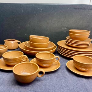 Könnte beinhalten: Eine Sammlung von hellbraunem Keramikgeschirr, darunter Tassen, Untertassen, Schalen und Teller. Das Set enthält einen Sahnekännchen und eine kleine Schüssel. Die Artikel sind auf einer blauen Oberfläche mit dunklem Hintergrund angeordnet.