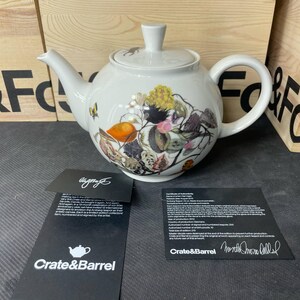 May include: A white ceramic teapot with a lid, featuring a colorful botanical and insect illustration on the body. Two black informational cards are placed in front, one with the Crate&Barrel logo and the other a certificate of authenticity.