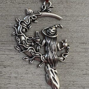 May include: Silver-toned pendant necklace featuring a witch figure within a crescent moon design. The witch is depicted with long hair and a pointed hat, holding a candle and a small orb. The moon is adorned with vines and leaves.