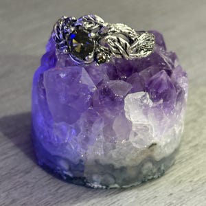 May include: A silver ring with a dark gemstone and intricate leaf design rests on a purple amethyst crystal. The ring's details are highlighted against the crystal's natural texture and color variations. The ring is a piece of jewelry.