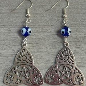 May include: A pair of silver-colored earrings. Each earring features a dangling, openwork design of a stylized trinity knot with a pentagram, crescent moons, and stars. Above the knot is a blue bead with a white eye design, suspended from a silver-colored hook.