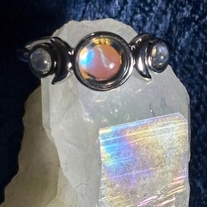 May include: A silver ring featuring a central iridescent stone, flanked by two crescent moon designs and smaller, round stones. The ring is set on a white crystal, with a dark blue background.