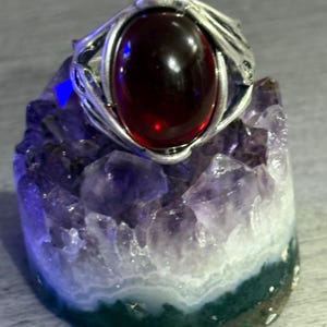 May include: A silver ring featuring a deep red, oval gemstone. The ring is displayed on a purple and white amethyst geode with a green base. The ring's design includes detailed, swirling metalwork around the gemstone.