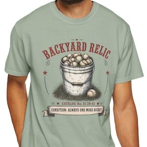 Op de afbeelding: Een saliegroen t-shirt met korte mouwen en een afbeelding van een emmer vol honkballen. De tekst luidt BACKYARD RELIC, CATALOG No. 11-28-41 en CONDITION: ALWAYS ONE MORE BUCKET. Het ontwerp bevat rode sterren en een banier-element.