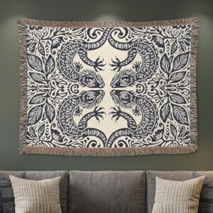 May include: A woven tapestry featuring a symmetrical design of four stylized lizards and foliage in navy blue on an off-white background. The tapestry is rectangular with a fringed edge and is displayed above a grey sofa.