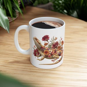 Puede incluir: Taza de café de cerámica blanca con una ilustración detallada de un dragón barbudo adornado con flores coloridas. La taza está llena de café oscuro y tiene un asa blanca. El diseño incluye tonos de rojo, naranja y amarillo.