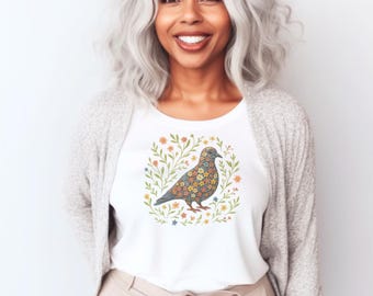 Könnte beinhalten: Weißes T-Shirt mit einem floralen Vogeldesign. Der Vogel ist mit bunten Blumen geschmückt und von einem Kranz aus grünen Blättern und kleinen Blumen umgeben. Die Person, die das T-Shirt trägt, hat graues Haar und eine hellgraue Strickjacke.