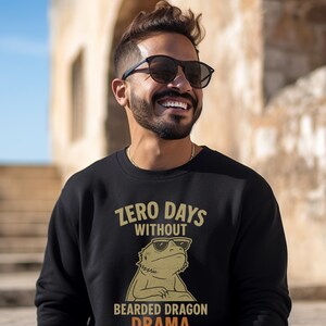 May include: Black sweatshirt with the text "ZERO DAYS WITHOUT BEARDED DRAGON DRAMA" and a graphic of a bearded dragon wearing sunglasses. The text and graphic are in shades of gold and orange.