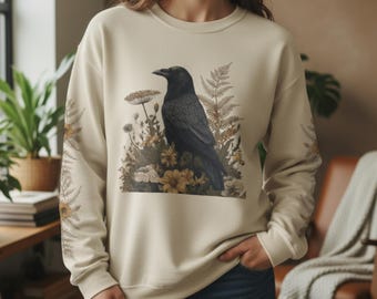 May include: Cream-colored sweatshirt featuring a detailed illustration of a black crow perched amidst wildflowers and ferns. The sleeves have a floral design. The design is centered on the front of the sweatshirt.