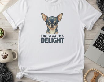 May include: White t-shirt featuring a watercolor illustration of a Chihuahua with the text "FIRST OF ALL I'M A DELIGHT" in dark blue. The dog has black and brown fur and large ears. The shirt is folded on a light wood surface.