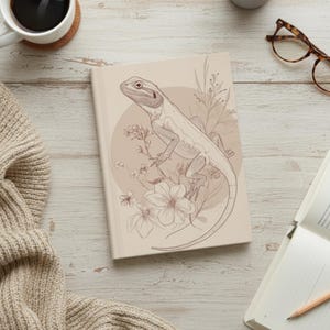 Puede incluir: Un diario beige con un dibujo lineal detallado de un dragón barbudo y elementos florales. El lagarto está representado en una rama, rodeado de flores y hojas. El diario descansa sobre una superficie clara con vetas de madera.