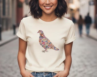 Könnte beinhalten: Cremefarbenes T-Shirt mit einem bunten Vogeldesign aus Blumen. Der Vogel ist in Lila-, Orange-, Gelb- und Blautönen gehalten. Das Shirt hat hochgekrempelte Ärmel und wird mit blauen Jeans kombiniert.