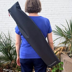 May include: A long, black carrying case with a red zip is carried over a person's shoulder. The case has the text "Song of Joy Studios" printed on the side. The person is wearing a blue top and navy trousers.