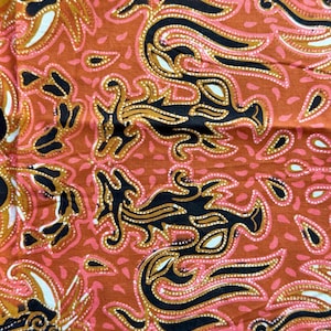 May include: Close-up of a fabric with a repeating pattern. The design features stylized bird-like figures in black and gold, set against a reddish-orange background. Pink and white accents add contrast to the design.