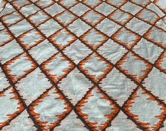 May include: Close-up of a fabric with a repeating diamond pattern. The diamonds are outlined in dark brown and filled with a light blue-gray color. The edges of the diamonds have an orange zig-zag design.