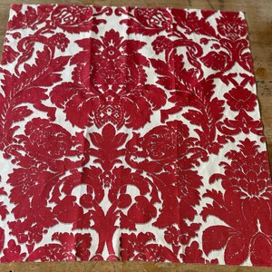 Op de afbeelding: Vierkante stof met een rood damastpatroon op een witte achtergrond. Het ontwerp bevat sierlijke bloemen- en bladerenmotieven, wat een klassieke, elegante uitstraling creëert. De stof wordt waarschijnlijk gebruikt voor woondecoratie of knutselprojecten.