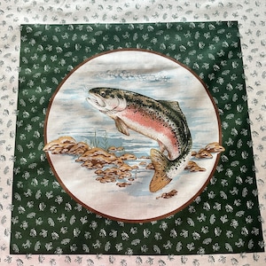 May include: A square fabric with a central circular design featuring a leaping rainbow trout. The trout has pink, red, and green hues. The background is a light blue with rocks and water. The border is dark green with a repeating pattern of small white insects.